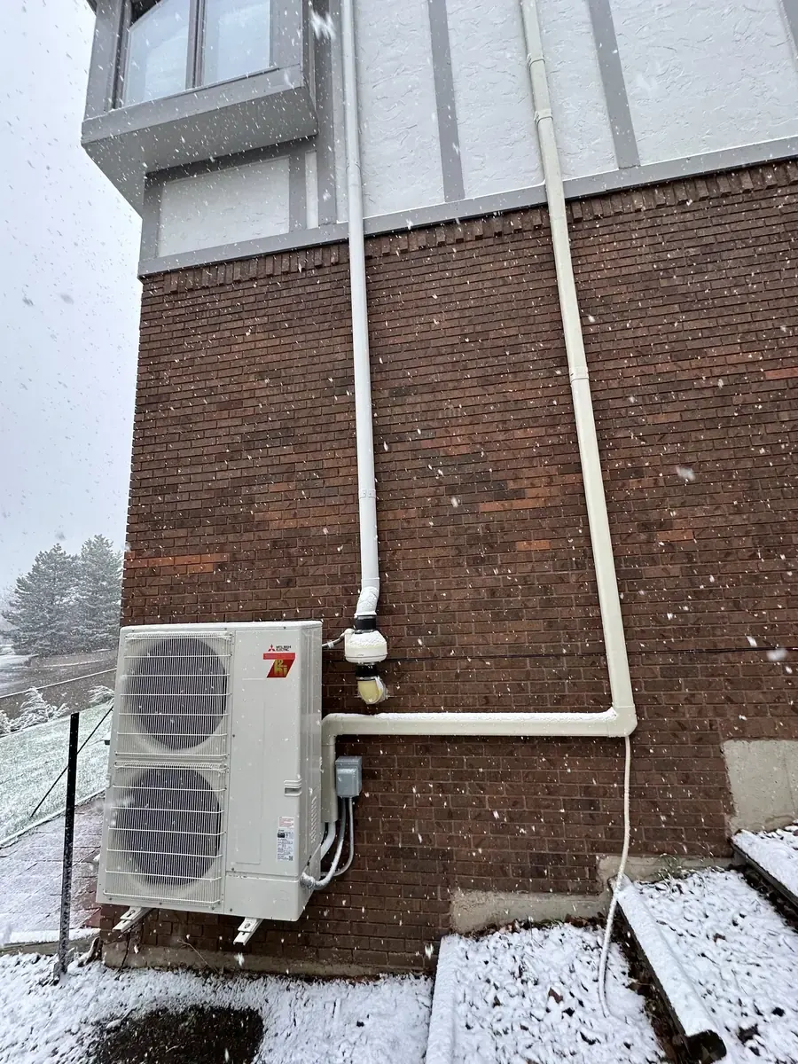 A cold-climate heat pump running in falling snow against a brick wall.